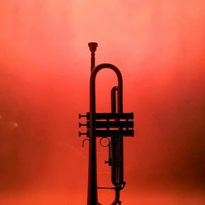 A single trumpet stands silhouetted against a vivid orange background, creating a dramatic musical image.